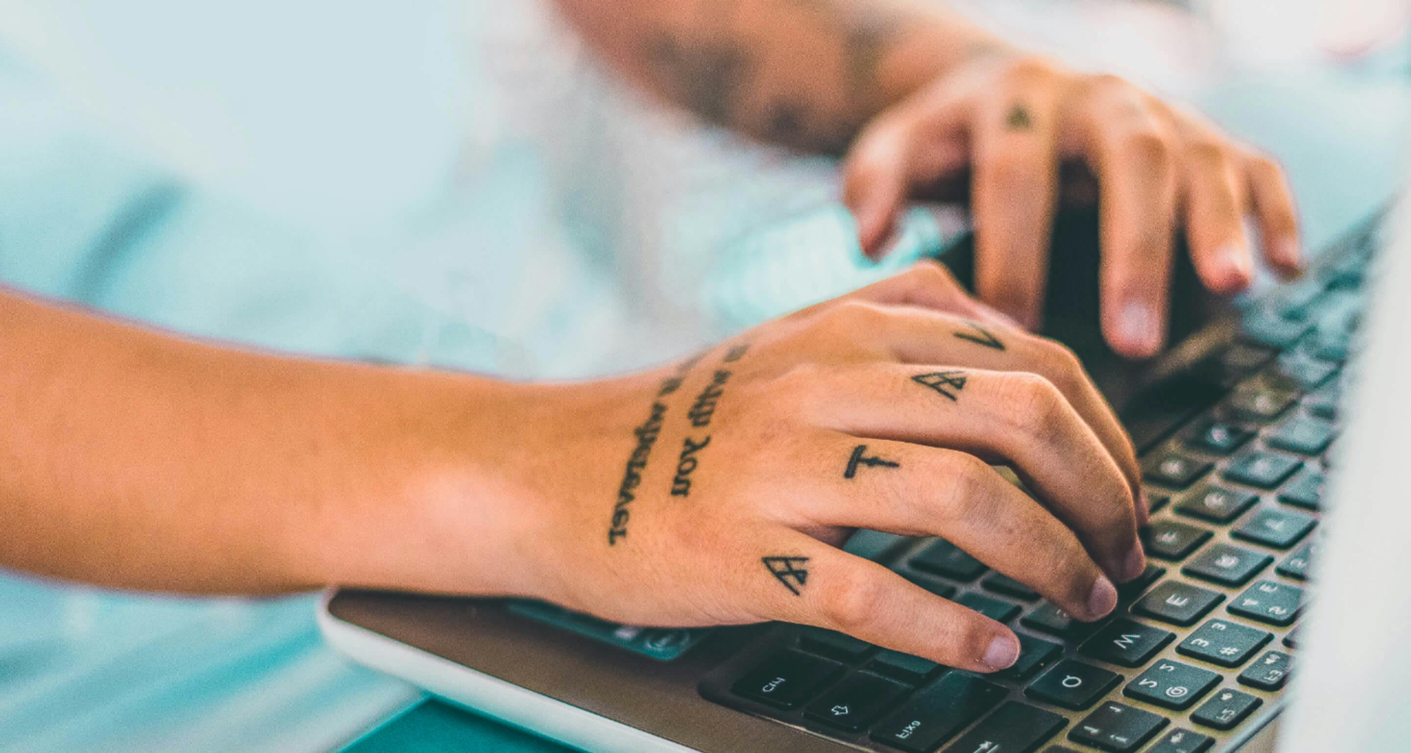 Mãos tatuadas digitando em um teclado de computador